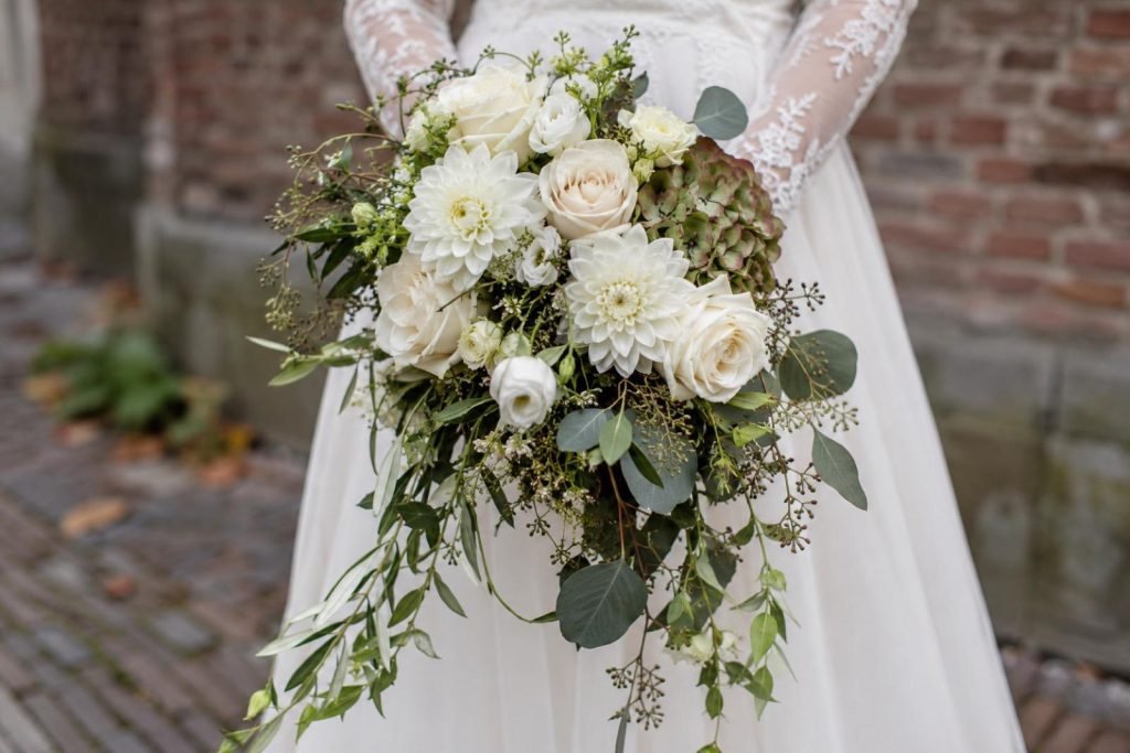 Bruidsboeket van witte rozen en dahlia’s, vastgehouden voor een witte jurk